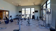 This fitness center offers a bright and well-appointed space designed to support a comprehensive workout routine. The room features a checkered tile floor that provides a durable, easy-to-clean surface, while two ceiling fans ensure consistent air circulation throughout the space. Large floor-to-ceiling windows and a glass door along one wall flood the interior with natural light and offer views of an outdoor pool area and palm trees, creating an inviting and motivating atmosphere. The soft blue-grey walls contribute to a calm and focused environment. Cardio equipment includes treadmills, ellipticals, upright and recumbent bikes, and a rowing machine, providing a wide variety of aerobic training options. Strength and resistance training is supported by a cable machine and dedicated weight benches. Two wall-mounted flat-screen televisions are positioned for easy viewing from multiple workout stations, enhancing the overall experience. The single-story layout keeps everything accessible and organized, with equipment thoughtfully arranged to allow for comfortable movement between stations. Overall, this fitness center combines functionality with a clean, modern aesthetic, making it a well-rounded amenity for regular use.