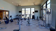 This fitness center offers a bright and well-appointed space designed to support a comprehensive workout routine. The room features a checkered tile floor that provides a durable, easy-to-clean surface, while two ceiling fans ensure consistent air circulation throughout the space. Large floor-to-ceiling windows and a glass door along one wall flood the interior with natural light and offer views of an outdoor pool area and palm trees, creating an inviting and motivating atmosphere. The soft blue-grey walls contribute to a calm and focused environment. Cardio equipment includes treadmills, ellipticals, upright and recumbent bikes, and a rowing machine, providing a wide variety of aerobic training options. Strength and resistance training is supported by a cable machine and dedicated weight benches. Two wall-mounted flat-screen televisions are positioned for easy viewing from multiple workout stations, enhancing the overall experience. The single-story layout keeps everything accessible and organized, with equipment thoughtfully arranged to allow for comfortable movement between stations. Overall, this fitness center combines functionality with a clean, modern aesthetic, making it a well-rounded amenity for regular use.