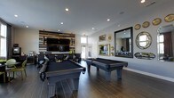 Located at 2980 Mark Andrew Dr in Dublin, Ohio, this single-floor amenity space is designed with both style and entertainment in mind. The centerpiece of the space is a spacious game room featuring two full-sized pool tables with sleek black felt, positioned on rich wood-look tile flooring that adds warmth and sophistication to the room. A striking reclaimed wood accent wall serves as a dramatic backdrop, anchoring a large wall-mounted flat-screen TV that makes the area ideal for both gaming and watching events. Recessed lighting illuminates the space evenly, complementing the modern, neutral-toned walls. An oversized decorative mirror framed in black adds depth and visual interest to one wall, accompanied by tasteful gold and sunburst wall accents that lend an upscale feel. Additional seating and a secondary mounted TV are visible in the background, suggesting a lounge or bar area perfect for social gatherings. Natural light filters in through windows, keeping the space bright and inviting. The overall ambiance is contemporary and polished, blending industrial and modern design elements to create a versatile, community-focused entertainment destination.