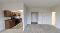 This single-floor residential space offers a practical and straightforward layout designed for comfortable everyday living. The living area is finished with wall-to-wall carpet, providing a warm and quiet underfoot feel, and flows openly toward the entryway, which features a clean white door and a louvered closet for convenient storage.

Adjacent to the living area, the kitchen is separated by a partial wall that maintains an open connection between the two spaces. The kitchen is equipped with warm wood-toned cabinetry, black appliances including a gas range, and a sink positioned beneath a skylight that draws natural light into the cooking area. The kitchen floor transitions to hardwood-style flooring, offering a functional and easy-to-maintain surface.

The neutral white walls throughout the space create a bright, clean atmosphere that lends itself well to personalization. The overall layout is efficient, with clearly defined zones for living and cooking. A thermostat panel is visible on the living room wall, and the louvered closet near the entry adds practical storage just steps from the front door. The space presents a well-organized, move-in-ready environment suited for a range of lifestyles.