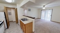 This single-floor unit is designed with a practical, open layout that connects the kitchen and living areas seamlessly. The kitchen is equipped with warm oak cabinetry, light-toned laminate countertops, a double-basin sink, a gas range, a dishwasher, and a refrigerator. Tile flooring runs throughout the kitchen and utility areas, while a peninsula with a dark countertop overhang serves as a breakfast bar, visually separating the kitchen from the carpeted living area.

The living room is generously sized, featuring a ceiling fan with integrated lighting and a sliding glass patio door dressed with vertical blinds, allowing natural light and outdoor access. A second window near the kitchen adds additional brightness to the shared space.

Two bedrooms are present, each carpeted and featuring windows that bring in natural light. Both rooms offer comfortable proportions suitable for standard furnishings.

The unit includes two bathrooms. The full bathroom features a tub-shower combination, toilet, single vanity with oak cabinetry, and a large mirror. The half bathroom provides a vanity and toilet with tile flooring. A utility area with an electrical panel is accessible from the kitchen corridor, adding functional convenience to the overall layout.