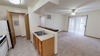 This single-floor unit is designed with a practical, open layout that connects the kitchen and living areas seamlessly. The kitchen is equipped with warm oak cabinetry, light-toned laminate countertops, a double-basin sink, a gas range, a dishwasher, and a refrigerator. Tile flooring runs throughout the kitchen and utility areas, while a peninsula with a dark countertop overhang serves as a breakfast bar, visually separating the kitchen from the carpeted living area.

The living room is generously sized, featuring a ceiling fan with integrated lighting and a sliding glass patio door dressed with vertical blinds, allowing natural light and outdoor access. A second window near the kitchen adds additional brightness to the shared space.

Two bedrooms are present, each carpeted and featuring windows that bring in natural light. Both rooms offer comfortable proportions suitable for standard furnishings.

The unit includes two bathrooms. The full bathroom features a tub-shower combination, toilet, single vanity with oak cabinetry, and a large mirror. The half bathroom provides a vanity and toilet with tile flooring. A utility area with an electrical panel is accessible from the kitchen corridor, adding functional convenience to the overall layout.