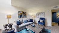 This two-floor residence offers a thoughtfully designed layout that balances comfort and functionality across its living spaces. The main level centers around a spacious living room highlighted by soaring vaulted ceilings and large windows that flood the room with natural light. Neutral-toned carpet anchors the space, while the open layout allows for generous furniture arrangements and an inviting ambiance.

The kitchen is efficiently arranged in a galley style, featuring white cabinetry with dark granite-style countertops, wood-look flooring, and black appliances including a range, refrigerator, and dishwasher. The clean, bright cabinetry provides ample storage, and the layout is optimized for ease of use.

Ascending to the upper level, the bedroom is tucked beneath a striking pitched roofline with exposed beams, creating an airy, loft-like atmosphere. A window brings in soft natural light, and the carpeted flooring adds warmth. A landing area at the top of the staircase provides additional functional space, complete with a dresser nook.

The bathroom features a wide vanity with white cabinetry, a large mirror, dark stone-style countertops, and a separate shower-tub combination, offering a clean and practical design throughout. Overall, the space presents a cohesive, light-filled environment with consistent neutral tones and modern-casual touches.