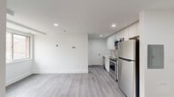 This efficiently designed 1-bedroom, 1-bathroom unit offers a thoughtful layout across a single floor, making smart use of every square foot. The open living and kitchen area is anchored by a contemporary kitchen featuring floor-to-ceiling white cabinetry, a tiled backsplash, light quartz countertops, and a full suite of stainless steel appliances including a refrigerator, electric range, built-in microwave, and dishwasher. Recessed lighting illuminates the space, complementing the light grey wood-style flooring that runs throughout. The living area is bright and clean with crisp white walls, providing a neutral canvas that feels open and airy. The bedroom is an intimate, well-defined space with a window offering an outdoor view, allowing natural light to fill the room. The bathroom features a full soaking tub with overhead shower, white mosaic tile flooring, and a frosted window that brings in soft natural light while maintaining privacy. The overall aesthetic is modern and minimalist, with consistent finishes, clean lines, and a neutral palette that creates a cohesive, move-in-ready feel throughout the unit.