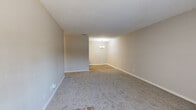This single-floor unit offers a well-organized layout designed for comfortable everyday living. The living area is generously proportioned, featuring plush wall-to-wall carpeting, neutral beige walls, and a textured ceiling throughout. A sliding glass door with vertical blinds allows natural light to fill the space and provides access to the outdoors. The living area flows seamlessly into a dedicated dining space, distinguished by warm hardwood-style flooring and an elegant chandelier overhead, creating a welcoming atmosphere for meals and gatherings. The galley-style kitchen is efficiently designed with ample cabinetry finished in a light gray tone with gold hardware, white laminate countertops, and white appliances including a range and refrigerator. A flush-mounted ceiling fixture provides bright, even lighting. The bedroom is a well-sized, carpeted retreat with a window dressed in white blinds that invites natural light. The bathroom features a clean, all-white tiled tub and shower combination, offering a simple and functional design. Clean lines, consistent neutral tones, and practical finishes are found throughout, making this a move-in-ready space with a cohesive, understated aesthetic.