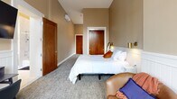 Located at 888 Delaware Ave in Buffalo, NY, this efficiently designed one-bedroom, one-bathroom unit offers a thoughtful layout across a single floor. The kitchenette area is positioned near the entrance and features white shaker-style cabinetry, a light countertop, a compact stainless steel refrigerator, and a sink — all illuminated by a large window that frames views of the surrounding brick architecture and fills the space with natural light.

The generously sized bedroom is the heart of the unit, boasting impressively tall ceilings that lend an open, airy feel well beyond what the footprint might suggest. Warm beige walls are complemented by classic white wainscoting, plush carpet flooring, and multiple windows that bring in abundant daylight and treetop views. Rich wood-toned doors add warmth, while brass wall-mounted sconces provide refined accent lighting. A comfortable seating area with leather-upholstered chairs and a small desk rounds out the bedroom's functionality.

The bathroom is well-equipped with a grey vanity, marble-look countertop, a large mirror with vanity lighting, and a full tub-shower combination adorned with elegant marble-style tile surround and built-in niche shelving. The overall aesthetic throughout the unit is clean, neutral, and modern with classic touches.