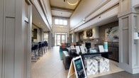 Located at 8718 Wintersweet Ln in Charlotte, NC, the Blakeney community clubhouse is a thoughtfully designed gathering space that blends sophisticated aesthetics with welcoming functionality. Upon entry, visitors are greeted by a grand foyer featuring a striking two-tone tile floor in a classic diamond pattern, setting an elegant tone for the rest of the space. Soaring double-height ceilings flood the main lounge with an impressive sense of openness, further enhanced by large transom windows that draw in abundant natural light.

The central lounge area is anchored by a handsome stone fireplace, flanked by wall-mounted artwork and decorative accents, creating a warm and inviting focal point. Comfortable leather seating and a long reception-style desk provide both a welcoming atmosphere and practical amenities for residents. Structural columns with detailed millwork and wainscoting throughout lend the space a refined, classic character.

Adjacent to the main lounge, a bar area with tall seating offers a casual gathering spot, while a separate dining nook with a round table and natural window light provides a quieter retreat. Glass-paneled doors lead directly to a fitness center, visible from the dining area, ensuring convenient access. The overall ambiance is polished and modern, with a cohesive neutral palette accented by teal and warm earth tones.