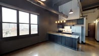 This single-floor urban loft showcases a striking blend of industrial and contemporary design. The open-concept layout centers around a well-appointed kitchen featuring sleek dark cabinetry that extends along the wall, complemented by light granite countertops that provide a bold contrast. Stainless steel appliances, including a French door refrigerator with ice maker, an electric range, and an over-the-range microwave, give the kitchen a polished, modern feel. A multi-light pendant fixture suspended above the countertop adds warm, focused illumination, enhancing the space's sophisticated ambiance.

One of the most distinctive architectural elements is the exposed industrial ductwork running across the dark ceiling, lending the space an authentic loft character. Polished concrete-style flooring flows throughout, adding to the urban aesthetic while remaining easy to maintain. A large picture window floods the space with natural light and frames an expansive city view, connecting the interior to the surrounding urban landscape. The dark wall tones create a dramatic backdrop that ties together the industrial and modern elements seamlessly, resulting in a cohesive, stylish living environment ideal for urban living.