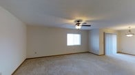 This single-floor space features a generously sized main living area finished with wall-to-wall neutral carpet, creating a clean and versatile foundation. The room is characterized by smooth, light-colored walls accented with classic wood baseboards, contributing to a simple and functional aesthetic. A ceiling fan with an integrated light fixture is centrally mounted, offering both air circulation and ambient lighting. A window along the far wall allows natural light to filter into the room, brightening the interior. The textured ceiling adds subtle dimension overhead. An adjacent area or hallway is visible to the right, suggesting additional connected spaces within the layout. Overall, the space presents a straightforward, move-in-ready environment well-suited for a variety of uses.