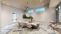 Located in Clermont, FL, this single-floor commercial lobby space offers a thoughtfully designed interior that blends modern aesthetics with a welcoming, functional atmosphere. The space is anchored by a striking patterned carpet in layered shades of gray, blue, and cream, which adds texture and visual depth throughout the open floor plan.

A central workstation and built-in bench seat a focal point of the space, crowned by a dramatic sculptural pendant light with an open, radial design that draws the eye upward toward the high clerestory windows. These elevated windows flood the interior with natural light, enhancing the airy, open feel of the room. Additional tall potted plants, including a prominent fiddle-leaf fig, bring organic warmth and life to the bright, neutral walls.

A bold teal shiplap accent wall anchors a dedicated lounge seating area, where round tables and upholstered chairs are arranged beneath a pair of framed abstract artworks and globe wall sconces, creating an inviting collaborative nook. On the opposite side, teal-painted shelving with built-in cabinetry provides functional storage with decorative detail.

Recessed lighting and a glass-paneled entry door complete the polished, professional atmosphere, making this a versatile and visually engaging space well-suited for client-facing or community use.