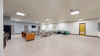 This well-appointed single-floor community space offers a generous, open layout designed to support a variety of activities and social interactions. The interior is bright and welcoming, illuminated by recessed flush-panel ceiling lights that cast an even, comfortable glow across light-toned vinyl plank flooring throughout.

The kitchen area is fully equipped with warm wood cabinetry, dark stone countertops, a full-size refrigerator, a dishwasher, and a double sink. A bold orange island with a granite-style countertop serves as both a prep area and a casual gathering point, with seating along one side. A coffee station adds additional functionality to this well-organized culinary space.

Adjacent to the kitchen, the open dining and lounge area accommodates multiple seating arrangements, including café-style tables with mixed chairs and teal upholstered booth seating along the walls. A wall-mounted television and colorful nature-themed artwork contribute to a lively yet relaxed ambiance. A cozy lounge nook with soft teal armchairs provides a quieter retreat.

On the opposite end of the space, a dedicated classroom area features two large chalkboards spanning the wall, flexible seating, and a distinctive yellow accent door, making it suitable for instruction, workshops, or group programming. The overall design blends functionality with a cheerful, community-focused aesthetic.