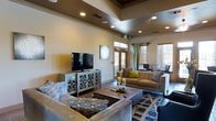 This single-floor space offers a well-appointed layout that blends functional amenities with inviting design. The kitchen is outfitted with dark cabinetry, stainless steel appliances including a full-size French door refrigerator, granite countertops, and recessed lighting overhead. A peninsula counter separates the kitchen from the adjacent recreation area, where a full-sized pool table provides an entertaining focal point. The transition between tile and hardwood-style flooring subtly defines the different zones within the space. Circular decorative mirrors accent the wall near the billiards area, adding a modern touch to the décor. The lounge area is a standout, featuring soaring tray ceilings with a rich, contrasting finish and an array of recessed lights that create a warm, layered ambiance. Large windows dressed with drapes and blinds allow natural light to fill the room, while French doors offer a connection to the exterior. The seating area is generously furnished with plush sofas, patterned accent chairs, and a dark wood coffee table anchored by a geometric area rug. A media console with a flat-screen television completes the lounge, making this space ideal for both relaxation and social gatherings.