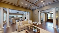 This single-story clubhouse is designed with an upscale, welcoming aesthetic that balances elegance with functionality. The main gathering area immediately draws attention with its striking coffered ceiling, accented by dark wood trim and recessed lighting that casts a warm, inviting glow throughout the space. Classic white columns define distinct zones within the open floor plan, adding architectural character while maintaining a seamless flow between areas.

Rich hardwood flooring extends throughout the primary living spaces, complementing the warm taupe wall tones that lend a cohesive, sophisticated feel. A cozy seating area anchored by a traditional fireplace with a marble surround and custom white mantle offers a refined focal point, flanked by oversized windows that allow generous natural light and views of the surrounding outdoor area.

The open-plan layout accommodates multiple seating arrangements, including plush upholstered sofas and accent chairs, making it well-suited for both intimate gatherings and larger community events. A fully equipped kitchen features granite countertops, white cabinetry, stainless steel appliances, and a peninsula with bar seating — ideal for catering and entertaining. The entry foyer, framed by a classic wood door, connects the various areas of the clubhouse with ease and purpose.