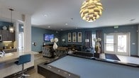 This single-floor amenity lounge is designed for both entertainment and relaxation, offering a well-appointed space with a cohesive, contemporary aesthetic. The room is anchored by a full-sized billiards table at its center, positioned beneath a stunning sculptural pendant light that serves as a bold focal point. Rich blue-gray walls lend a sophisticated ambiance throughout the space, complementing the warm, layered furnishings.

Multiple seating arrangements are thoughtfully distributed across the lounge, including plush sofas and accent chairs upholstered in complementary textures, providing ample space for gathering. A wall-mounted flat-screen television is positioned at one end of the room, creating a dedicated media area. Decorative framed artwork and ornamental floor lamps add to the polished, curated feel of the interior.

Recessed ceiling lighting works in tandem with the statement pendant and floor lamps to ensure the space is well-lit and inviting. Natural light filters in through large windows dressed with patterned drapes, offering a connection to the outdoors. Decorative vases and floral arrangements are placed throughout, adding organic texture and visual interest. Overall, this lounge delivers a dynamic, multifunctional environment well-suited for social gatherings and leisure activities.