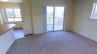This single-floor space features a carpeted room finished in neutral, light tones that lend a calm and airy atmosphere. The room is anchored by a sliding glass door fitted with vertical blinds, which opens directly onto a private balcony enclosed by a metal railing. The balcony offers elevated views of surrounding evergreen trees, bringing a natural backdrop and ample natural light into the interior. The wall-to-wall carpet is a muted beige tone, contributing to the room's understated and practical aesthetic. A paneled interior door on the left side of the room provides access to an adjoining space with hardwood-style flooring visible through the doorway, suggesting a varied flooring layout throughout the unit. The walls are painted in a soft, warm off-white shade, complementing the neutral palette of the space. Light switches and outlets are positioned conveniently along the walls. The overall design is straightforward and functional, with the sliding glass door and balcony serving as the standout feature, offering a strong connection to the outdoors and enhancing the sense of openness within the room.