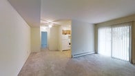 This single-floor residence offers a straightforward, functional layout designed for comfortable everyday living. The main living area is generously sized, featuring neutral wall-to-wall carpeting that adds warmth underfoot. A ceiling fan provides both lighting and air circulation, enhancing the room's comfort year-round. A baseboard heating unit runs along the exterior wall, ensuring efficient climate control throughout the colder months.

Sliding glass doors line one side of the living area, fitted with vertical blinds that allow for adjustable natural light while providing access to an outdoor space. The open sightlines connect the living area to the kitchen, which is equipped with wood-toned cabinetry, a white refrigerator, and a white range with overhead ventilation — offering a clean, practical cooking environment.

A hallway extends from the main living space, leading to additional rooms visible in the background, including what appears to be a room with blue-toned flooring. The overall color palette is neutral throughout, with light beige walls and consistent flooring, creating a cohesive and move-in-ready environment.
