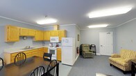 This single-floor unit offers a practical and well-organized layout designed to meet everyday living needs. The kitchen is equipped with light oak cabinetry providing ample storage, white appliances including a refrigerator, electric range, and built-in microwave, as well as a double sink and counter space for food preparation. Crown molding lines the ceiling throughout, adding a clean, finished look to the interior. Bright fluorescent lighting ensures the space is well-lit at all times. Adjacent to the kitchen, a dining area accommodates a dark-toned table and chair set, suitable for casual meals. The living area transitions seamlessly from the dining space and features neutral gray carpeting, creating a comfortable and cohesive atmosphere. A cushioned recliner chair and additional seating provide a relaxed setting for everyday use. The subdued gray wall tones throughout contribute to a calm, neutral ambiance. A standard interior door leads to an additional room, suggesting further private living quarters. Overall, the space is thoughtfully arranged to balance functionality with comfortable residential living.