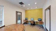 The business center is a thoughtfully designed single-level workspace that combines functionality with an inviting atmosphere. The room's most striking visual element is a bold yellow accent wall adorned with a subtle botanical pattern, creating an energetic focal point that adds personality to the otherwise neutral space. Recessed lighting in the white ceiling provides consistent, even illumination throughout the room. Along the accent wall, a continuous built-in desk surface accommodates three individual computer workstations, each equipped with a desktop monitor and ergonomic rolling chair in warm brown tones. This dedicated row of workstations is ideal for residents or guests needing reliable computer access. A rustic wooden round table positioned near the glass-paneled entry door offers an additional area for meetings or collaborative work. The door features a privacy shade and provides views to the exterior, allowing natural light to filter into the space. Neutral carpeted flooring and clean wall finishes contribute to a professional yet comfortable ambiance, making this business center a practical and well-appointed resource.
