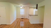 This single-floor residence offers a clean, open layout designed for comfortable everyday living. The main living area is anchored by gleaming hardwood floors that run throughout, reflecting natural light and lending warmth to the space. A ceiling fan with integrated lighting is centrally positioned overhead, providing both comfort and illumination. Vertical blinds dress the windows, offering privacy while still allowing ample light to fill the room.

A half-wall peninsula with a granite-style countertop separates the living area from the kitchen, creating a natural transition between the two spaces while maintaining an open, connected feel. The kitchen area features recessed lighting and dark cabinetry overhead, providing a functional and well-lit workspace. The peninsula also offers casual seating or serving potential, making it a practical focal point.

The walls are finished in a neutral, light tone that enhances the brightness of the space, while classic white baseboards frame the room cleanly. A radiator beneath one of the windows provides efficient heating. Overall, the space presents a straightforward, well-maintained interior with timeless hardwood flooring, practical amenities, and an inviting open-plan design suitable for a variety of living arrangements.