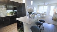 This single-floor residence offers a thoughtfully designed open-concept layout that effortlessly connects the kitchen, dining, and living areas into one cohesive and welcoming space. The kitchen is a standout feature, showcasing rich dark cabinetry that provides a bold contrast against the light granite countertops. A central island with a waterfall-edge granite surface serves as both a functional workspace and a casual dining spot, accommodated by modern wire-frame bar stools. Pendant lighting above the island adds a contemporary touch, while full-sized black appliances lend a sleek, uniform aesthetic. Built-in open shelving within the cabinetry offers convenient display and storage space. Light hardwood flooring flows throughout the space, enhancing the sense of continuity and warmth. The adjoining living area is bright and airy, benefiting from large windows that invite generous natural light. A ceiling fan adds to the comfort of the space. The bedroom, visible in the background, continues the open and light-filled atmosphere. Overall, the space strikes a balance between modern design and comfortable livability, making it both visually appealing and highly functional.
