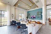 This thoughtfully designed single-floor community clubhouse offers a generous, open-concept layout that seamlessly connects a variety of functional and social spaces. Soaring double-height ceilings create an impressive sense of volume throughout, while large floor-to-ceiling windows and glass doors flood the interior with abundant natural light and provide direct views of the outdoor pool and surrounding courtyard.

The main gathering area features a sophisticated lounge zone anchored by a plush sectional sofa, leather ottomans, and a teal accent wall adorned with industrial-style shelving and a wall-mounted flat-screen TV. An ornate coffered ceiling with gold geometric detailing adds a refined architectural focal point to this seating area. Adjacent to the lounge, a formal dining space accommodates a large wooden dining table with high-backed chairs, illuminated by stylish pendant lighting and flanked by towering fluted columns.

A dedicated workstation area offers bar-height seating along a long communal desk, ideal for productivity or casual gatherings. The kitchenette zone is equipped with warm oak cabinetry, white quartz countertops, stainless steel appliances, a commercial-grade coffee station, and open floating shelves. Large-format stone tile flooring flows consistently throughout, unifying the space with a clean, modern aesthetic. The overall ambiance is warm, contemporary, and inviting.