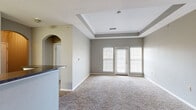 Located at 516 Stone Dr in Manhattan, Kansas, this single-floor unit offers a well-designed layout with comfortable living spaces throughout. The living room is a standout feature, showcasing an elegant tray ceiling that adds architectural depth and visual height to the room. Soft, neutral-toned carpet covers the floor, while large windows fitted with blinds and a set of glass double doors allow abundant natural light to fill the space. The open flow connects the living area to the kitchen via a stylish arched passageway. The kitchen is well-appointed with warm wood cabinetry, dark granite-style countertops, tile flooring, and a full suite of white appliances including a refrigerator, range, and dishwasher. A breakfast bar adds additional counter space and casual dining convenience. The bedroom features the same neutral carpet and grey walls as the living area, with dual windows providing good natural light. The bathroom is finished with tile flooring, a wood vanity with dark countertop, and a full tub-shower combination. Consistent grey wall tones and white trim throughout the unit create a cohesive, clean aesthetic that feels move-in ready.