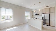 Located on Market Street in Suffolk, Virginia, this well-appointed one-bedroom, one-bathroom residence offers a thoughtfully designed layout across a single floor. The open-concept living and kitchen area serves as the heart of the unit, where natural light pours through multiple double-hung windows framing views of the surrounding cityscape. The kitchen is a standout feature, showcasing gray shaker-style cabinetry, a herringbone-pattern tile backsplash, stainless steel appliances including a refrigerator, microwave, and range, and a large white quartz island with an undermount sink. Pendant lighting above the island adds a contemporary touch. Light-toned flooring flows consistently throughout the space, enhancing the clean, modern aesthetic. A hallway with a louvered-door closet connects the main living area to the private bedroom, which is a comfortable size with a bright window overlooking the city below. The bedroom also provides direct access to a spacious walk-in closet equipped with wire shelving. The bathroom features a white vanity with warm-toned countertop, gold-accented fixtures, a large mirror, and a glass-enclosed shower, combining functionality with refined style.