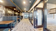 Located at 2256 Fox Valley Center in Aurora, Illinois, this single-floor commercial space offers a thoughtfully curated interior that balances comfort with contemporary design. The dining area is defined by a row of high-back upholstered booths finished in a muted teal fabric, each paired with warm wood-toned tables and separated by sleek metal dividers for a sense of semi-private dining. A striking nature-inspired wallcovering featuring silhouetted winter trees runs along the back wall, complemented by Edison-style wall sconces that add a warm, intimate glow to each booth. The ceiling is finished in a deep teal tone with wood trim accents, adding visual depth and a cohesive color palette throughout. Underfoot, an abstract-patterned carpet in neutral tones with dark brushstroke detailing ties the space together. Adjacent to the main dining area, glass-paneled private rooms enclosed by dark-framed doors offer flexible meeting or consultation spaces, with curated artwork and a distinctive round teal accent table visible in one room. At the far end, a glass-walled fitness area is visible through large windows, providing connectivity between the lounge and recreational amenities. Natural light filters in through exterior-facing windows, enhancing the overall ambiance of this well-appointed space.