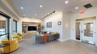 Located at 1211 East 7th Street in Tulsa, Oklahoma, this single-floor community amenity space combines contemporary design with a warm, functional layout. Polished concrete floors run throughout the main area, anchoring a cohesive modern aesthetic accented by track lighting and recessed fixtures overhead.

The central lounge area serves as the heart of the space, featuring a built-in light wood entertainment wall with open shelving and a large flat-screen television. Comfortable seating — including a sectional sofa, a wooden coffee table, and a pair of bold yellow accent chairs — creates an inviting gathering environment. A triptych city map of Tulsa adds a locally inspired decorative touch to the walls.

Adjacent to the lounge, a dining and social zone offers a tall communal table with bar stools beneath pendant lighting, alongside smaller round dining tables with mid-century modern chairs. A dedicated kitchenette is equipped with light wood cabinetry, dark countertops, a stainless steel refrigerator, a sink, and additional appliances, finished with a graphic patterned accent wall.

Large floor-to-ceiling windows and glass openings along one side of the space flood the interior with natural light and provide direct views of a covered outdoor patio. The patio, framed by exposed brick and mature trees beyond a wrought iron fence, offers additional seating for outdoor enjoyment. The overall ambiance is clean, contemporary, and community-focused.