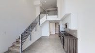 This two-story space showcases a clean, modern design characterized by crisp white walls and an open, airy atmosphere throughout. The main level features polished concrete flooring that extends through the living area and into the kitchen, contributing to the industrial-modern aesthetic. The kitchen is thoughtfully appointed with wood-toned cabinetry, dark countertops, a tile backsplash, and stainless steel appliances including a range, microwave, and refrigerator. A recessed door at the rear of the main level leads to an additional space, offering functional utility.

The staircase, constructed with a steel frame and horizontal metal railing, ascends prominently and serves as a visual focal point, complementing the contemporary character of the space. Soaring double-height ceilings on the main floor amplify the sense of openness and volume.

The upper level transitions to warm wood-look plank flooring, creating a contrasting and inviting environment. A skylight on the upper floor introduces natural light, brightening the space considerably. The upper level's open layout provides a versatile area suitable for various uses, overlooking the main floor below through the open stairwell.
