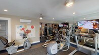 The fitness room at 550 Los Arbolitos in Oceanside is a well-appointed exercise facility designed to support a range of workout routines. The single-level space features a combination of flooring materials, including blue rubberized flooring along the perimeter and modular carpet tiles in the central workout zone, creating defined areas for different activities.

The cardio section is equipped with multiple treadmills, elliptical trainers, and stationary bikes, including both upright and recumbent models. A multi-station strength machine anchors one end of the room, complemented by a large motivational athletic mural that adds visual energy to the space. Full-length mirrors along one wall expand the sense of space and allow users to monitor their form during exercise. A wall-mounted flat-screen television provides entertainment during workouts.

Natural light fills the room through a generous row of windows and a set of glass-paneled double doors that open to a landscaped outdoor deck area, offering pleasant views of the surrounding greenery. Recessed lighting and a ceiling fan maintain a comfortable atmosphere. A wall-mounted mini-split HVAC unit ensures climate control year-round. The facility also includes restroom access and a defibrillator unit, reflecting a commitment to user safety and convenience.