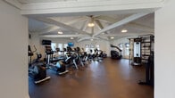 Located at 934 Shorepoint Ct in Alameda, California, this two-floor fitness center is a comprehensively outfitted facility designed to support a full range of workout routines. The upper floor is distinguished by its striking vaulted ceiling with exposed white cross-beam trusses, creating an airy, spacious atmosphere. This level houses an extensive cardio area featuring multiple treadmills, elliptical machines, and stair climbers, all arranged to take advantage of the natural light streaming through surrounding windows. Wall-mounted televisions are positioned throughout for entertainment during workouts. A dedicated treadmill room on this floor provides additional focused cardio space, also equipped with mounted screens. A functional area with a punching bag, TRX suspension system, medicine balls, kettlebells, and stability equipment offers versatile training options. A lounge nook with bold orange accent chairs and access to an outdoor balcony adds a relaxing transition space. The lower floor expands the strength training offerings, featuring multiple cable machines, a power rack, a comprehensive dumbbell rack, weight benches, and large floor-to-ceiling mirrors that visually extend the space. An air bike and additional multi-station cable systems round out the equipment on this level. Dark rubber flooring throughout both floors provides durability and safety, while white walls and recessed lighting maintain a clean, modern environment. A staircase connects both levels seamlessly.