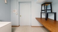 Located at 1700 E Main St in Kent, Ohio, this shared laundry room is a compact, single-floor utility space designed for practical, everyday use. The room is constructed with painted concrete block walls in a light blue tone, giving it a clean and utilitarian appearance. Flooring consists of neutral, low-pile carpet that extends throughout the space.

The laundry area is equipped with two top-loading washing machines and two dryers, positioned along adjacent walls to maximize the use of the available floor space. Each washer-dryer pair is neatly arranged and connected to dedicated hookups, with a flexible aluminum exhaust duct channeling dryer ventilation through the wall. A standalone water heater is also present in the room, situated in the corner near the dryers.

A wood-laminate folding table provides a convenient surface for sorting and folding laundry, making the room more functional for users. A standard interior door with a gold-tone knob serves as the entry point, accompanied by a light switch and posted informational signage on the adjacent wall. The overall layout is straightforward and efficient, offering all essential laundry amenities within a well-organized, no-frills space.