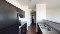 This single-floor residence offers a thoughtfully designed layout centered around a sleek galley-style kitchen. The kitchen is outfitted with rich, dark wood cabinetry that extends to the ceiling, accented by elegant crown molding that adds a refined architectural touch. Granite-look countertops provide generous prep space on both sides of the kitchen, while the stainless steel double basin sink offers practicality and style. Black appliances, including a refrigerator, electric range with overhead microwave, and additional cooking appliances, create a cohesive and modern aesthetic. Track lighting mounted on the ceiling provides focused illumination throughout the cooking space. The flooring throughout is a deep, rich hardwood that adds warmth and sophistication to the interior. Crown molding details are carried consistently through the space, lending a polished, upscale feel. A hallway visible from the kitchen leads further into the residence, hinting at additional living areas beyond. The overall ambiance is modern yet inviting, with a neutral color palette of crisp white walls contrasting beautifully against the dark cabinetry and flooring to create a space that feels both stylish and functional.