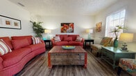 This single-floor unit offers a well-organized layout with distinct living, dining, kitchen, and sleeping areas, all connected by durable wood-style flooring throughout the main spaces. The living room serves as the heart of the unit, featuring textured ceilings, chair-rail walling, and bold red sofas centered around a rustic wooden trunk coffee table. Colorful artwork and teal accent lamps contribute to the space's vibrant, eclectic character. A wide entryway hall leads into the living area, with additional art displayed along the walls.

The dining area connects seamlessly to the living room and is highlighted by a large sliding glass door that floods the space with natural light and provides access to an outdoor area with views of surrounding greenery. A dark wood dining table with Windsor-style chairs accommodates multiple guests comfortably.

The kitchen features warm oak cabinetry, ample counter space, a window for natural light, and full appliances including a dishwasher, range, and refrigerator. A floral area rug adds a decorative touch.

The primary bedroom offers carpeted flooring, a king-sized bed setup, and natural light from a window with blinds. The second bedroom, also carpeted, accommodates two twin beds with coastal-inspired décor. The bathroom includes a single vanity, mirror, and a shower-tub combination.