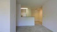 This single-floor space features a straightforward, open-concept layout that connects the kitchen and living area in a cohesive, functional arrangement. The kitchen is positioned behind a half-wall peninsula with a laminate countertop, creating a natural division between the cooking and living spaces while maintaining an open sightline. Within the kitchen, white cabinetry lines the upper walls, and a full-size refrigerator is visible alongside standard appliances. A fluorescent ceiling light fixture illuminates the kitchen area, while a track lighting fixture serves the adjacent living space, allowing for directional light control. The living area is finished with wall-to-wall carpeting in a neutral beige tone, contributing to a warm, understated ambiance. Walls throughout are painted in a clean white, enhancing the sense of brightness in the space. The overall design is minimal and utilitarian, with no ornate detailing, making it well-suited for straightforward, comfortable living. The open flow between the kitchen and living area maximizes the usability of the available space.