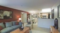 This single-floor residence showcases an open-concept design that seamlessly connects the living room, dining area, and kitchen. The living room is anchored by a plush sofa in a muted blue-gray tone, complemented by a rustic wooden coffee table and a cozy armchair with accent pillows. A bold terracotta-red accent wall adds warmth and visual contrast to the otherwise neutral color palette of soft whites and beige tones.

The dining area sits adjacent to the living space, furnished with a round table and upholstered chairs beneath a stylish pendant light fixture. Wall art adds a decorative touch, enhancing the refined yet approachable atmosphere. The kitchen is partially separated by a breakfast bar with dark countertops, offering a functional divide while maintaining the open feel. Light wood cabinetry and modern appliances are visible from the main living area.

Plush wall-to-wall carpeting runs throughout the main living spaces, contributing to the unit's warm and comfortable feel. The layout is thoughtfully arranged to maximize flow between areas, making the space feel both functional and inviting for everyday living and entertaining.
