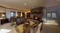 This single-floor space showcases a warm, craftsman-inspired design defined by rich dark wood trim, exposed beam accents, and neutral-toned walls throughout. The main lounge area serves as a central gathering space, anchored by a striking floor-to-ceiling stone fireplace with an active hearth. Plush leather seating, a patterned area rug, and built-in wooden shelving flanking the fireplace contribute to the room's inviting, lodge-like ambiance. Recessed lighting complements the warm tones of the space.

Adjacent to the lounge, a reception-style sitting area features elegant ironwork accent tables, a cushioned armchair, and dual windows fitted with horizontal blinds that frame outdoor views of the surrounding neighborhood and open landscape. Natural light filters generously across the carpeted floors throughout the day.

A wide corridor connects the main areas, decorated with wall-mounted botanical accents, framed artwork, and decorative barrels, lending an artistic and organic character to the transitional spaces.

The billiards room is a well-appointed recreation area centered around a full-size pool table beneath a pendant light fixture. Surrounded on multiple sides by large windows offering expansive exterior views, the room is bright and spacious, with additional seating stools and ample room for gameplay.