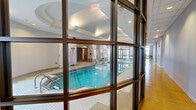 Located at 890 S State St in Chicago's South Loop neighborhood, this amenity floor offers a comprehensive wellness facility combining a fully equipped fitness center with an indoor aquatic area. The layout is thoughtfully organized, with distinct zones connected by light-filled corridors featuring warm bamboo hardwood flooring and large floor-to-ceiling glass partitions that provide visual continuity throughout the space.

The fitness center is generously proportioned and outfitted with a wide array of cardio equipment, including treadmills with individual TV monitors, elliptical machines, and stationary bikes. A dedicated strength training area houses a multi-station cable machine, free weights, medicine balls, and stability equipment. The gym features a drop ceiling with recessed lighting, rubber flooring, large windows admitting natural light, wall-mounted mirrors, and framed Chicago-themed artwork.

The indoor pool area is a standout feature, enclosed beneath an elegantly coffered ceiling with architectural detailing and soft ambient lighting. The lap pool is bordered by white tile and accessible via stainless steel railings. Glass wall partitions separate the pool from the surrounding corridor, maintaining an open, airy atmosphere. A hot tub is also present within the pool room. The entry corridor is accented with decorative greenery, clean white walls, and ample natural light from exterior-facing windows.