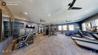 This fitness center offers a thoughtfully designed workout space that blends industrial character with modern functionality. The room is defined by a striking contrast of exposed brick walls and deep navy-blue painted walls, creating a dynamic and motivating atmosphere. Recessed lighting and multiple ceiling fans ensure the space remains well-lit and comfortable during workouts.

Natural light pours in through several large, multi-pane windows along the walls, offering views of surrounding greenery and enhancing the open, airy feel of the space. Full-length mirrors with gold-toned frames are mounted along the walls, adding both practicality and a refined aesthetic touch.

The fitness center is comprehensively equipped for a full-body workout. Cardio offerings include multiple treadmills and stationary bikes, while the strength training area features a multi-functional cable machine, a squat rack, a full dumbbell rack with a wide range of weights, adjustable benches, and various other resistance machines. Kettlebells and accessories are also available for functional training.

The flooring is covered with durable, cushioned carpet throughout, providing comfort underfoot. Glass-paneled double doors provide easy entry and exit, and a clearly marked emergency exit sign ensures safety. Overall, this fitness center is a well-appointed space suited for a comprehensive range of exercise routines.