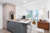 This single-floor unit delivers a thoughtfully designed living experience with a clean, contemporary aesthetic throughout. The living area serves as the centerpiece of the space, anchored by expansive floor-to-ceiling windows that frame sweeping urban skyline views, flooding the room with natural light. Sheer curtains complement the brightness while adding a soft, refined touch to the space.

The kitchen is outfitted with modern slate-toned cabinetry, stainless steel appliances including a Bosch range, and light stone countertops, offering both style and functionality. Light hardwood flooring runs throughout, reinforcing the space's cohesive, modern character.

The bedroom is a calm retreat, featuring a textured feature wall in warm neutral tones, a upholstered headboard, and a window with city views. A dedicated walk-in closet with built-in shelving and double hanging rods provides generous storage and organization.

The bathroom maintains the space's crisp aesthetic with an all-white palette, a wide vanity with an integrated sink, a large mirror, and a full tub-shower combination with tile surround. A hallway connects the rooms smoothly, accented by colorful wall art that adds a welcoming touch to the overall ambiance.