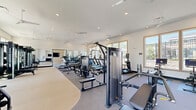 Located at 15576 Voyage Pl in Waldorf, MD, this fitness center offers a generously sized, single-floor exercise facility designed to support a full range of workout routines. The space is thoughtfully organized into distinct functional zones, providing ample room for both cardiovascular training and strength work without feeling crowded.

The cardio area features treadmills, ellipticals, and stationary bikes, including interactive cycling bikes, all positioned near large, wood-framed windows that invite abundant natural light and offer pleasant outdoor views. Ceiling fans overhead contribute to a comfortable workout environment throughout.

The strength training section is well-appointed with a full rack system, weight benches, a dumbbell rack, and a selection of cable and resistance machines, complemented by floor-length mirrors that visually expand the space and allow for form monitoring. The flooring transitions between carpet and durable exercise matting, suited to each activity zone.

A distinct studio area, finished with warm slatted wood wall paneling and crown molding details, provides a refined aesthetic and open floor space ideal for stretching, functional training, or group exercise. A wall-mounted television and a mirror-paneled fitness door add modern convenience to this section.

Recessed lighting and multiple ceiling fans throughout ensure the entire facility is bright, well-ventilated, and inviting for daily use.