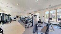 Located at 15576 Voyage Pl in Waldorf, MD, this fitness center offers a generously sized, single-floor exercise facility designed to support a full range of workout routines. The space is thoughtfully organized into distinct functional zones, providing ample room for both cardiovascular training and strength work without feeling crowded.

The cardio area features treadmills, ellipticals, and stationary bikes, including interactive cycling bikes, all positioned near large, wood-framed windows that invite abundant natural light and offer pleasant outdoor views. Ceiling fans overhead contribute to a comfortable workout environment throughout.

The strength training section is well-appointed with a full rack system, weight benches, a dumbbell rack, and a selection of cable and resistance machines, complemented by floor-length mirrors that visually expand the space and allow for form monitoring. The flooring transitions between carpet and durable exercise matting, suited to each activity zone.

A distinct studio area, finished with warm slatted wood wall paneling and crown molding details, provides a refined aesthetic and open floor space ideal for stretching, functional training, or group exercise. A wall-mounted television and a mirror-paneled fitness door add modern convenience to this section.

Recessed lighting and multiple ceiling fans throughout ensure the entire facility is bright, well-ventilated, and inviting for daily use.