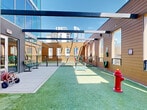 This dedicated outdoor dog park, located at 550 E Brooklyn Village Ave in Charlotte's Brooklyn Village neighborhood, offers a thoughtfully designed space for pets in an urban environment. The area is surfaced with artificial turf, providing a clean and comfortable space for dogs to play and exercise. Overhead steel beam pergola structures span the open-air space, offering partial shade while maintaining an open, airy feel beneath the Charlotte skyline.

The park is divided into distinct zones, including a larger open turf run and a smaller secondary turf area, separated by black metal fencing with a gated entry to ensure pet safety. Agility and play elements are incorporated throughout, including a jump bar obstacle and a classic red fire hydrant prop, adding both functionality and character to the space.

Along the perimeter, warm wood-toned siding panels and large windows allow natural light to filter through while framing views of the surrounding downtown buildings. Pet waste stations are conveniently mounted along the walls, and dog-themed benches provide seating for owners. Large glass doors connect the park to an adjacent interior space, making access seamless. The combination of practical amenities, playful design, and vibrant city views makes this a standout urban dog park.