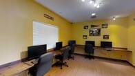 This single-floor computer lab is a functional and energetic workspace designed to accommodate multiple users simultaneously. The room is immediately distinguished by its bold yellow walls, which create a bright and stimulating atmosphere. A row of desktop computer workstations lines the perimeter of the room, each accompanied by ergonomic rolling chairs, providing ample space for focused individual work. The workstations are arranged along continuous desk surfaces that wrap around the room, maximizing available workspace.

Natural light filters in through a large window fitted with white horizontal blinds, complementing the overhead track lighting and recessed fixtures that ensure the space is well-lit throughout. The flooring consists of warm-toned hardwood-style planks, adding a clean and polished look to the room.

Wall décor celebrates Texas State University's Bobcat spirit, featuring a "Bobcat Dr." sign, a Texas State pennant, and several framed black-and-white photographs of Bobcat wildlife and athletic imagery. These elements give the lab a distinct sense of community identity and school pride.

Overall, the space balances practicality with personality — offering a well-equipped, welcoming environment for computer-based work, study, or research within a spirited academic setting.