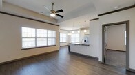 This single-floor residence showcases a thoughtfully designed open-concept layout that connects the living area and kitchen in one cohesive, flowing space. Rich dark hardwood-style flooring runs throughout, grounding the space with warmth and continuity. The living area benefits from generous natural light streaming through multiple large windows dressed with white horizontal blinds, creating a bright and welcoming atmosphere. A ceiling fan with a classic brass finish is centrally mounted within a tray ceiling detail, adding both function and architectural interest overhead. Crown molding accents the upper walls, lending a refined, polished character to the interior. The kitchen is positioned toward the rear and features a peninsula with dark countertops and a contrasting white upper cabinet arrangement. Stainless steel appliances, including a French-door refrigerator, add a contemporary touch, while a series of pendant lights hang elegantly above the peninsula, providing focused task lighting. The overall ambiance is clean, modern, and move-in ready, with neutral wall tones that complement the darker flooring and cabinetry. The open sightlines between spaces make the interior feel expansive, airy, and well-suited for both everyday living and entertaining.