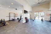 This single-story fitness center is designed to support a comprehensive workout experience within a bright, open-concept layout. The space is anchored by dramatically vaulted ceilings accented with crown molding, recessed lighting, and ceiling fans that keep the environment comfortable during intense sessions. The flooring transitions between warm hardwood near the entry and durable rubber tile throughout the main workout areas, clearly delineating zones for different training activities.

The entry area features wall-mounted cubbies and bench seating, providing a convenient spot to store belongings before exercising. Beyond the entryway, the main floor opens into a generously sized strength and functional training zone, equipped with a power rack, barbells, a dumbbell rack, weight bench, stability balls, and medicine balls. Motivational phrases such as "Hustle for Muscle" and "Harder. Better. Faster. Stronger." are displayed prominently on the walls throughout.

A dedicated cardio section hosts treadmills, an elliptical, and a rowing machine. Large floor-to-ceiling windows and sliding glass doors flood the interior with natural light and offer views of an outdoor pool area. Wall-mounted televisions and split-system air conditioning units enhance comfort. A mirrored wall and resistance training accessories complete the space, supporting a diverse range of fitness routines.