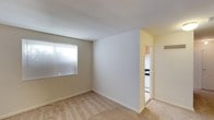 This single-floor space presents a straightforward and functional layout designed for comfortable everyday living. The main living area features wall-to-wall carpeting in a neutral beige tone, contributing to a warm and understated ambiance. Walls are finished in a clean white and light beige palette, creating a bright and open feel throughout. A window fitted with horizontal blinds allows natural light to filter into the living space while maintaining privacy. The room's simple design provides a versatile canvas for a variety of furniture arrangements and décor styles. An open doorway connects the living area to an adjacent kitchen, which is partially visible and features upper cabinetry in a warm wood tone alongside what appears to be standard appliances, including an oven range. The transition between spaces is smooth, with the carpeted flooring giving way near the kitchen entry. White baseboards frame the room cleanly, adding a crisp finishing detail. Overall, the space maintains a clean, move-in-ready aesthetic with a practical and efficient floor plan suited to a range of occupants.
