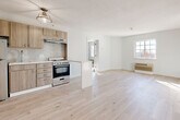 Located at 1151 E Main St in Meriden, Connecticut, this thoughtfully renovated single-floor unit offers a clean, modern aesthetic throughout. The open-concept living and kitchen area is anchored by light-toned hardwood flooring that extends seamlessly across the space, amplifying the bright, airy atmosphere created by natural light streaming through gridded windows. The kitchen is efficiently designed with warm wood-toned cabinetry, a tiled backsplash, white countertops, a stainless steel refrigerator, and a gas range with overhead ventilation — all oriented around a compact peninsula that defines the transition to the living area. The living space is generously proportioned, with neutral walls and a wall-mounted HVAC unit. A carpeted bedroom features its own window with outdoor views and a classic paneled door, offering a comfortable, quiet retreat. An additional room with hardwood floors and a double-door closet provides flexible use as a second bedroom or office. The bathroom is a standout feature, finished with large-format dark marble-style floor tiles, sculptural white wall tiles, a soaking tub, and a floating dark wood vanity with backlit mirror — delivering a spa-like contrast to the otherwise light and minimal palette of the unit.