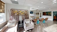 This single-floor clubhouse is thoughtfully designed to provide a welcoming and functional community space. The interior is anchored by an open-plan layout with wood-look plank flooring throughout, creating a warm and cohesive aesthetic. A drop ceiling with recessed lighting ensures the space is well-lit, complemented by abundant natural light streaming through large, white-framed windows that frame views of mature trees and manicured landscaping.

The main lounge area is generously appointed with a mix of seating options, including plush upholstered sofas, contemporary swivel chairs in warm cognac tones, and accent stools in a bold turquoise — adding a vibrant pop of color against the neutral palette. A large area rug grounds the central seating arrangement, while decorative elements such as abstract wall art, tall branch arrangements, and oversized potted plants add visual texture and depth.

A dedicated media area features a wall-mounted flat-screen television and a live-edge wood console table, making it a functional focal point for residents. Adjacent to this, a sunlit dining area provides a natural wood table with upholstered chairs, perfectly positioned beside floor-to-ceiling windows that overlook the outdoor greenery. The space balances comfort with contemporary design, making it a versatile and inviting environment for both social events and everyday use.